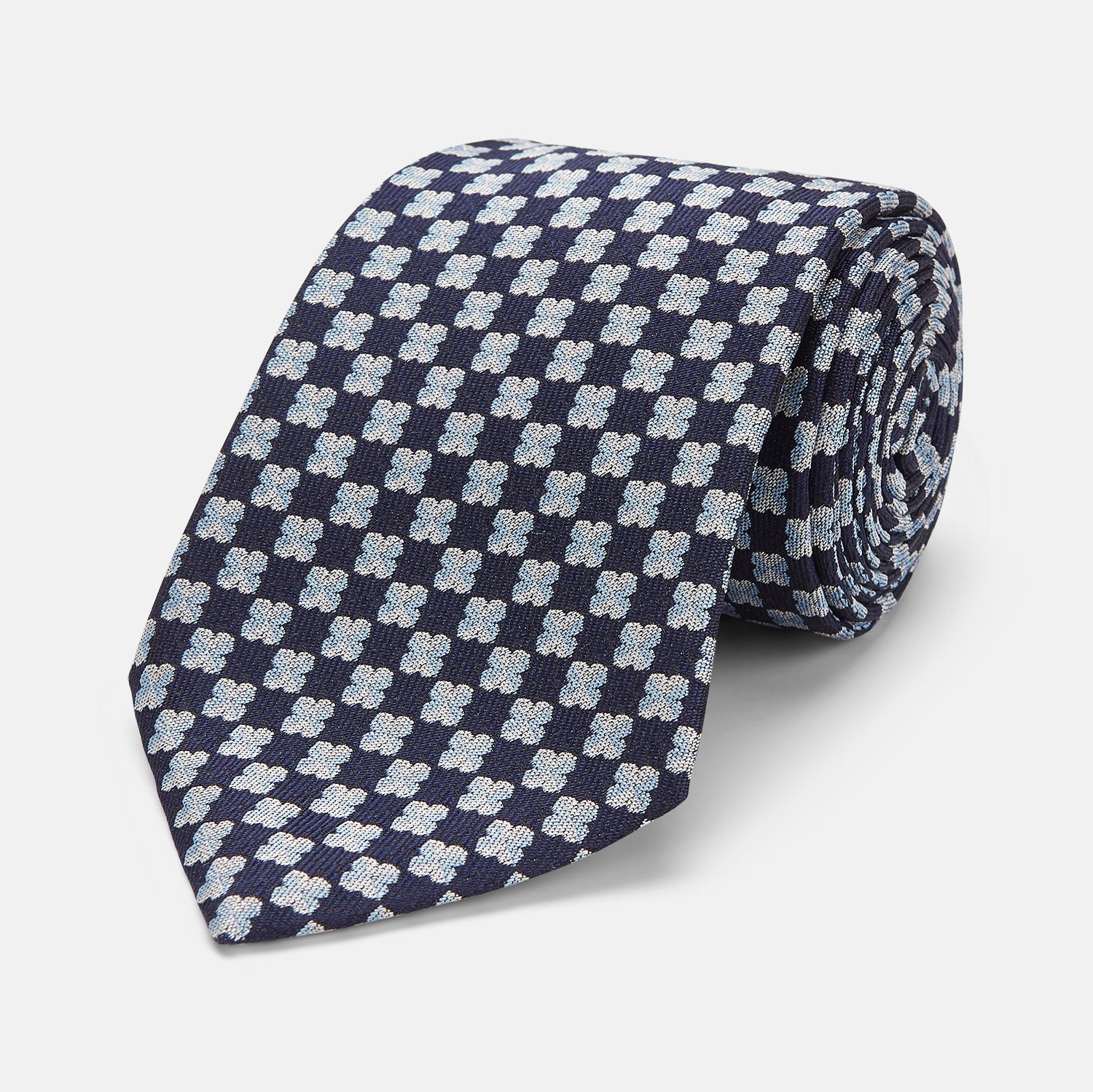View 1 of product The Turnbull & Asser Indigo Graphic Floral Silk Jacquard Tie features a repeating pattern of light blue and white geometric shapes, shown rolled up on a plain white background.