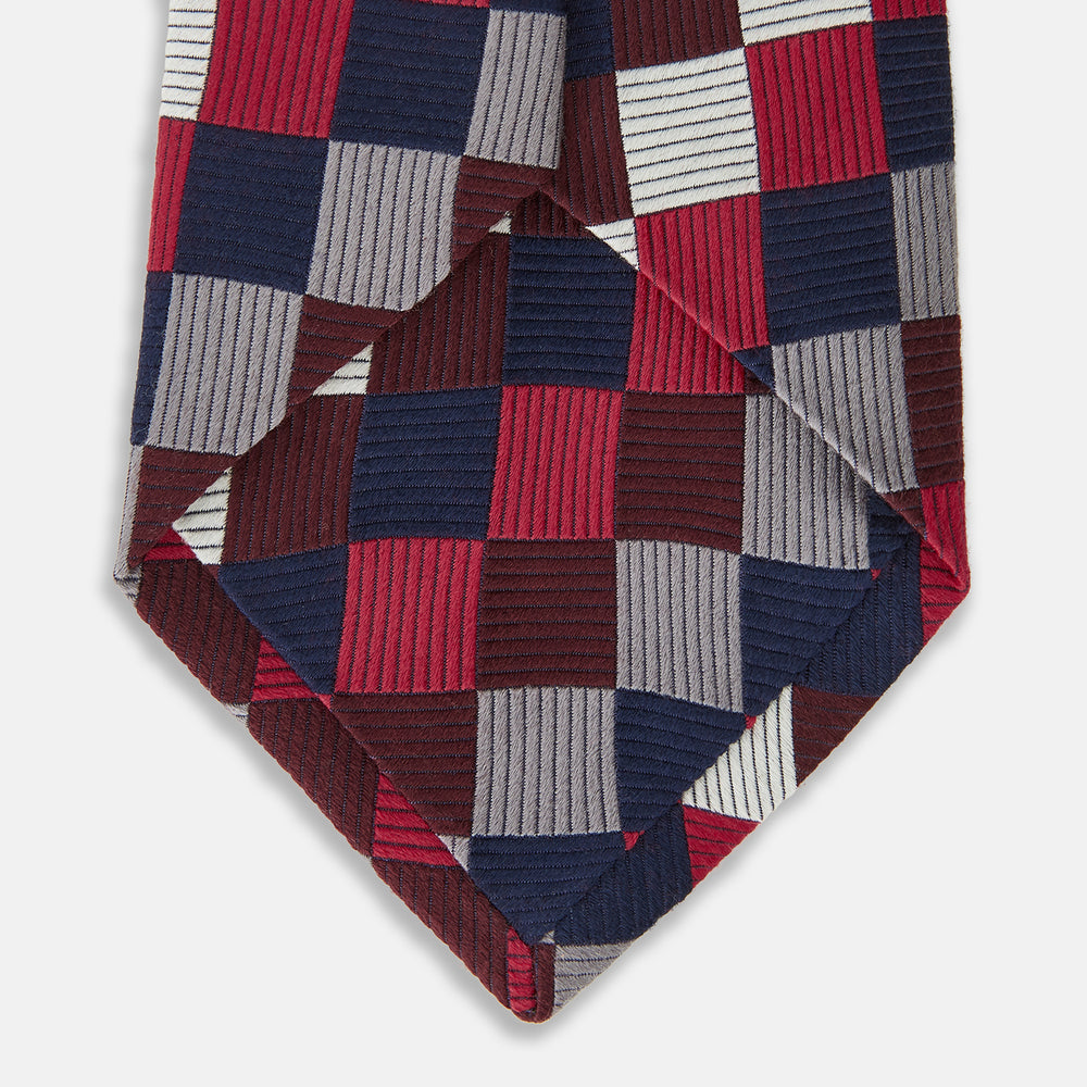 The Burgundy Checkerboard Silk Jacquard Tie by Turnbull & Asser features a bold autumnal pattern in red, navy blue, brown, gray, burgundy, and white squares, shown rolled on a plain white background.