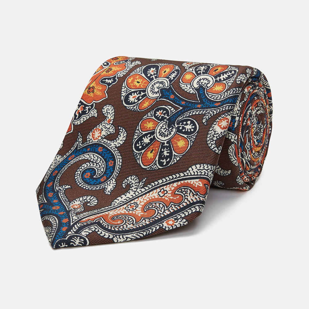 The Turnbull & Asser Chocolate and Orange Twisted Paisley Silk Tie for men showcases a vibrant orange, blue, and white paisley pattern inspired by classic Macclesfield designs, rolled on a plain white background.