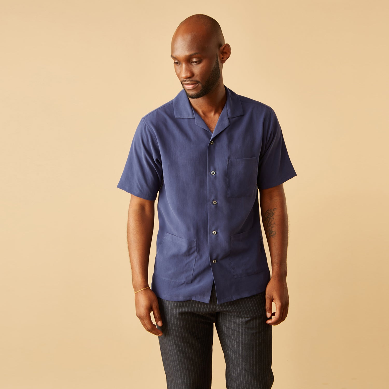View 1 of product A man stands against a beige backdrop, wearing the Turnbull & Asser Navy Holiday Fit Silk Phillips Shirt with Revere Collar and dark pinstripe pants. He gazes downwards with a relaxed expression, embodying effortless summer style.