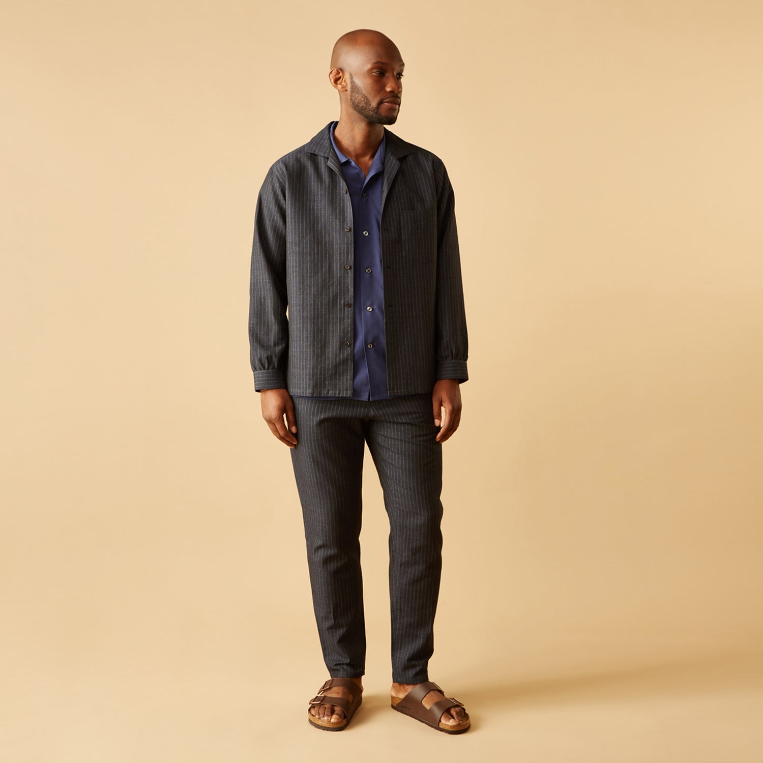 View 4 of product A man stands against a beige background, wearing a white t-shirt, Turnbull & Asser Navy Tailored Pinstriped Drawstring Trousers, and brown sandals. One hand is in his pocket as he looks straight at the camera.