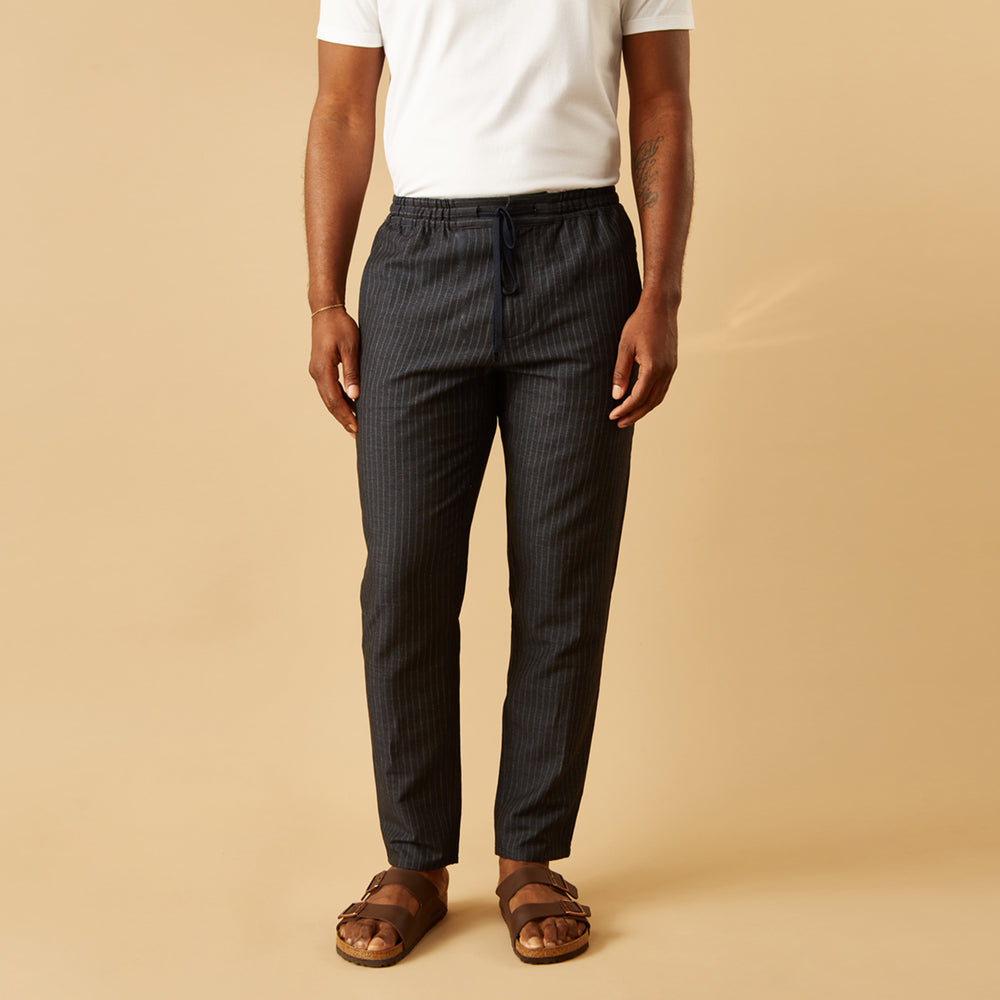 A man stands against a beige background, wearing a white t-shirt, Turnbull & Asser Navy Tailored Pinstriped Drawstring Trousers, and brown sandals. One hand is in his pocket as he looks straight at the camera.