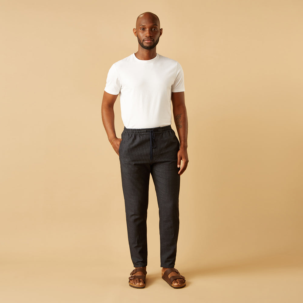 A man stands against a beige background, wearing a white t-shirt, Turnbull & Asser Navy Tailored Pinstriped Drawstring Trousers, and brown sandals. One hand is in his pocket as he looks straight at the camera.