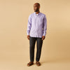 View 1 of product A man stands against a beige background wearing the Turnbull & Asser Lilac Weekend Fit Linen Blake Shirt with Stand Collar and 1-Button Cuffs, paired with dark pinstriped pants and brown sandals—a relaxed weekend look.