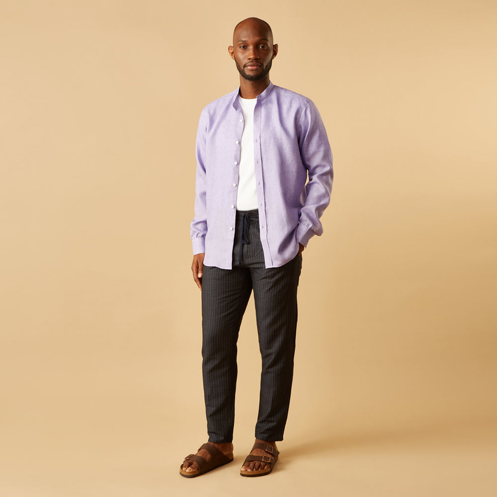 A man stands against a beige background wearing the Turnbull & Asser Lilac Weekend Fit Linen Blake Shirt with Stand Collar and 1-Button Cuffs, paired with dark pinstriped pants and brown sandals—a relaxed weekend look.