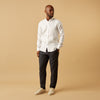 View 1 of product A man stands against a beige background, wearing the Turnbull & Asser White Linen Weekend Fit Blake Shirt, dark trousers, and beige sneakers. With one hand in his pocket and a neutral expression, he captures effortless weekend style.