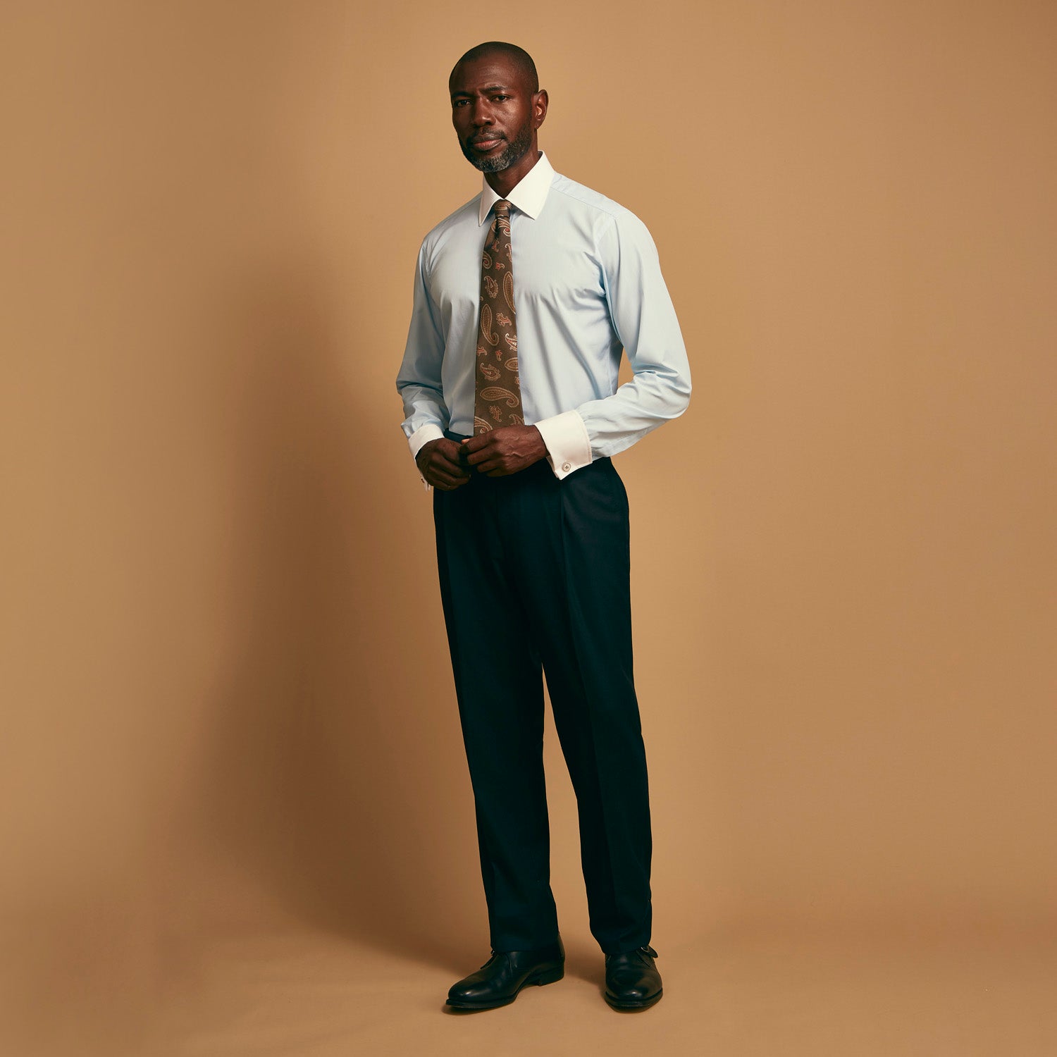 View 1 of product A man stands against a tan background wearing the Turnbull & Asser Mid Blue Micro-Check Regular Fit Shirt with White T&A Collar and Double Cuffs, paired with a brown patterned tie, dark trousers, and black dress shoes.