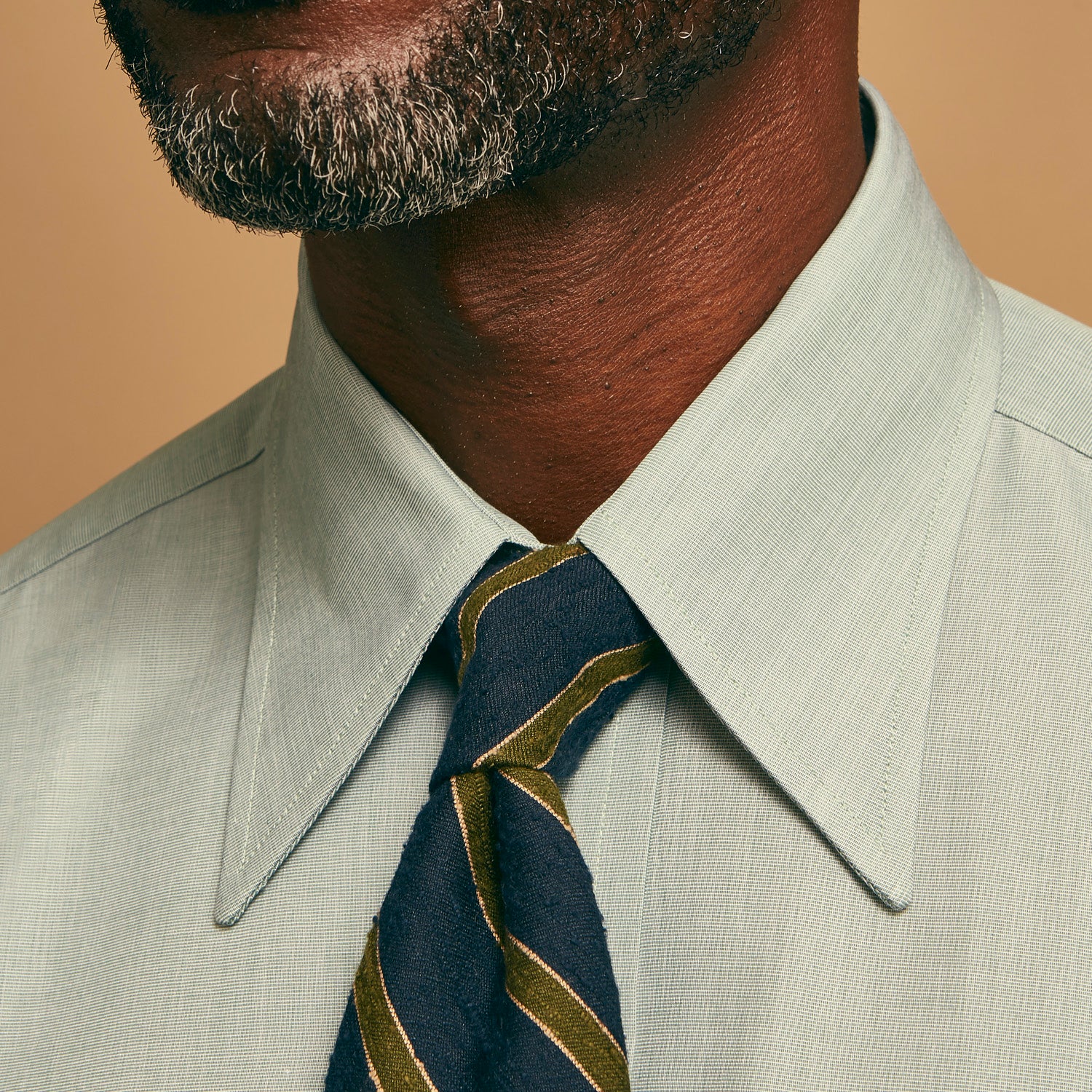 View 4 of product A man stands against a beige background wearing a Turnbull & Asser Green Weekend Fit Shirt with Long Point Collar and Single Button Cuffs, paired with a striped tie, dark trousers, and black dress shoes, hands in pockets and a neutral expression.