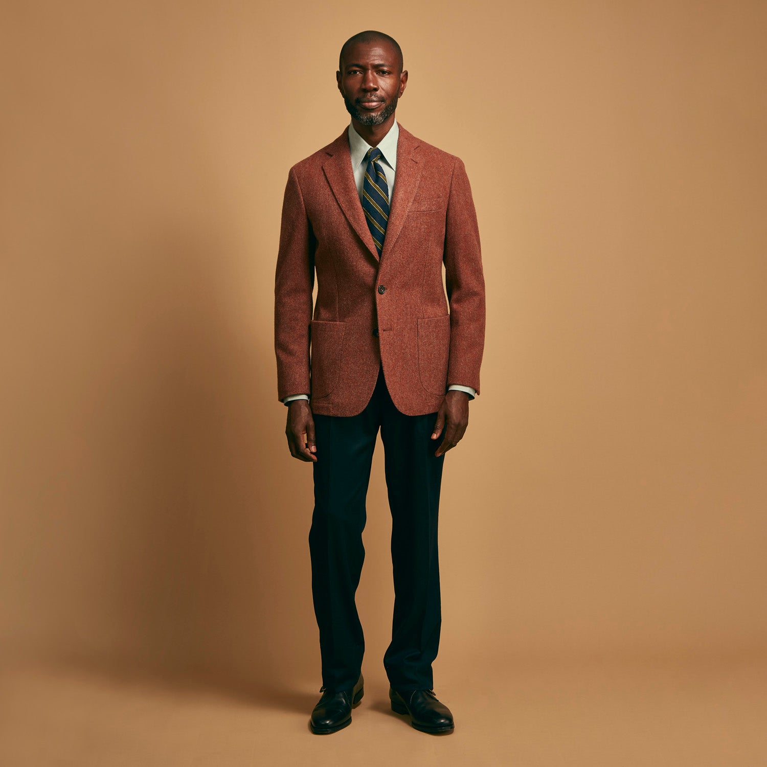 View 8 of product A man stands against a beige background wearing a Turnbull & Asser Green Weekend Fit Shirt with Long Point Collar and Single Button Cuffs, paired with a striped tie, dark trousers, and black dress shoes, hands in pockets and a neutral expression.
