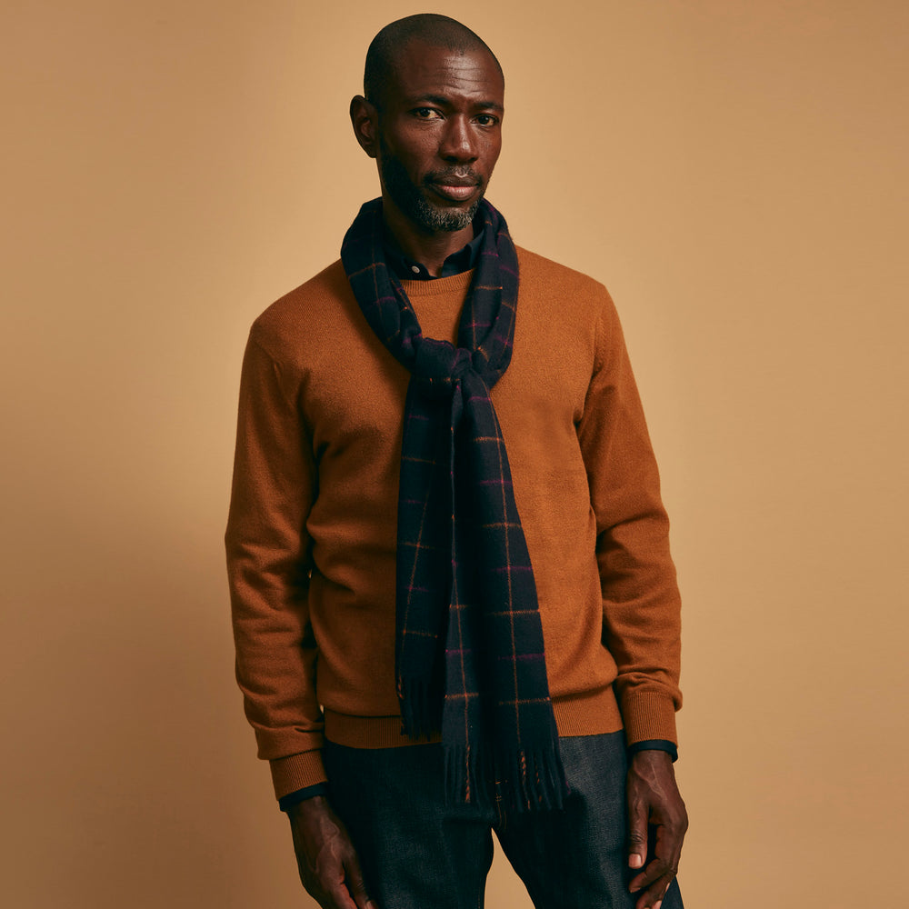A man stands against a beige background wearing a rust sweater, dark pants, and a Turnbull & Asser Purple Multi Tattersall Check Cashmere Scarf, looking confidently at the camera with a neutral expression.
