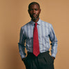 View 1 of product A man stands against a plain tan background wearing Turnbull & Asser's Blue Multi Chambray Stripe Weekend Fit Shirt with a Derby collar and 1 button cuffs, paired with a red patterned tie and gray trousers. His hands are in his pockets.