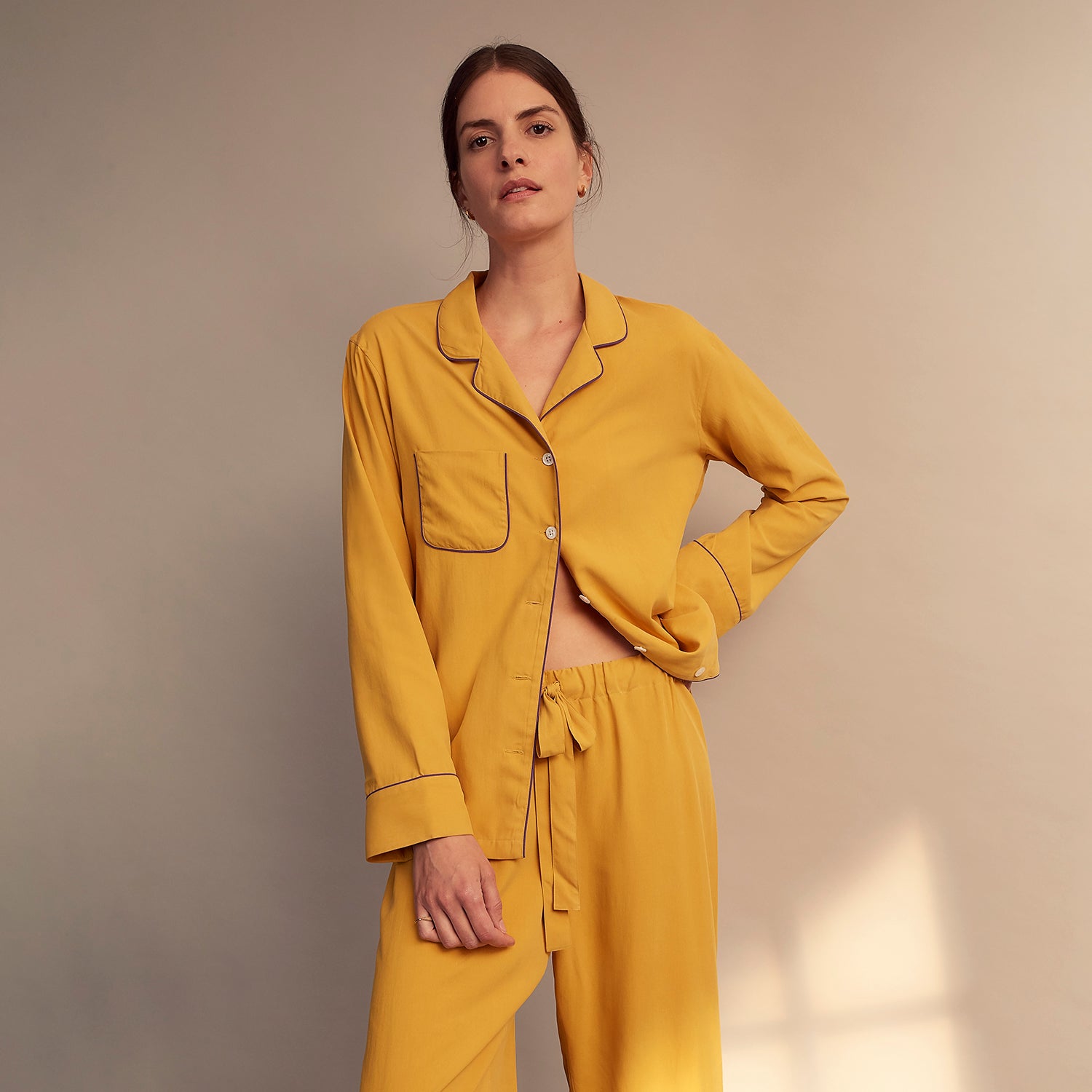 View 3 of product A woman stands barefoot on a gray floor wearing the Turnbull & Asser Women's Gold Silk Harriet Pyjamas Set, featuring a loose-fitting button-up top and long pants, against a plain beige background with soft light.