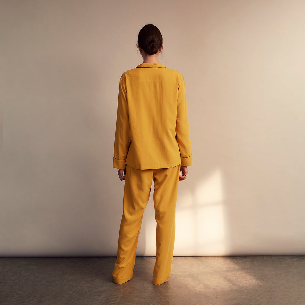 A woman stands barefoot on a gray floor wearing the Turnbull & Asser Women's Gold Silk Harriet Pyjamas Set, featuring a loose-fitting button-up top and long pants, against a plain beige background with soft light.