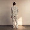 View 8 of product A person stands barefoot on a concrete floor, wearing the Turnbull & Asser Black & White Stripe Harriet Women’s Pyjama Set, featuring loose-fitting white pajamas with black vertical stripes, against a plain beige wall in soft side lighting.