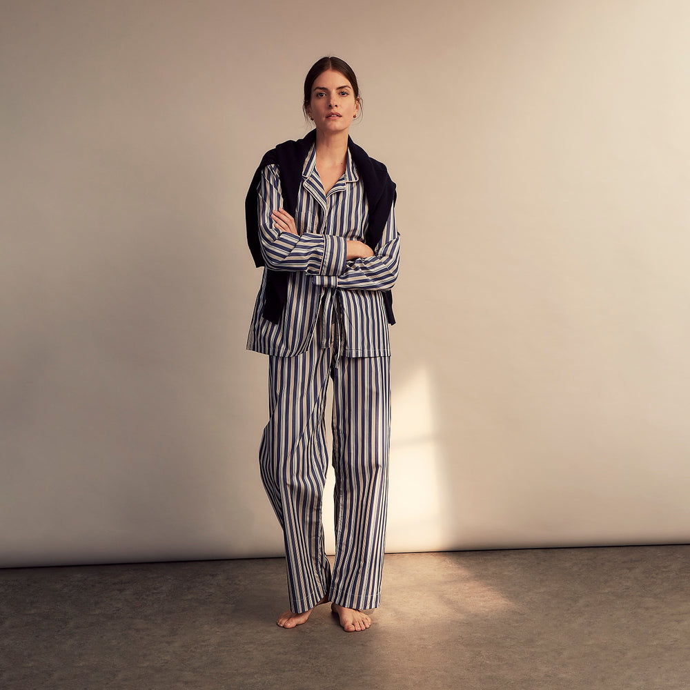 A person stands barefoot on a gray floor in the Turnbull & Asser Blue Multi Stripe Harriet Women’s Pyjama Set with a dark sweater over their shoulders—premium cotton pyjamas that make a perfect gift for her—against a plain beige background.