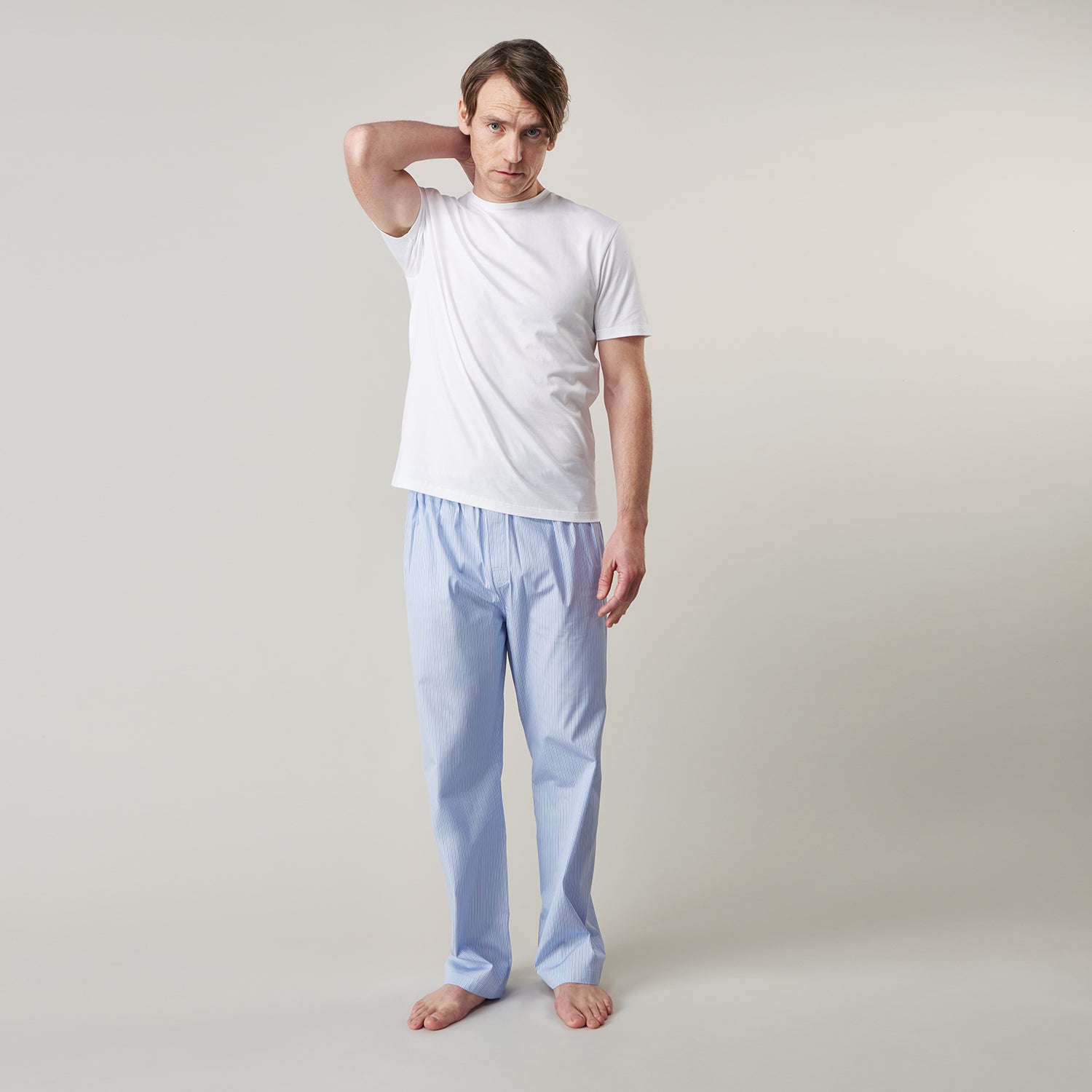 View 6 of product A man stands barefoot against a plain background, wearing a white short-sleeve T-shirt and Turnbull & Asser Blue Stripe Cotton Pyjama Trousers in a contemporary fit, looking straight ahead with a neutral expression.