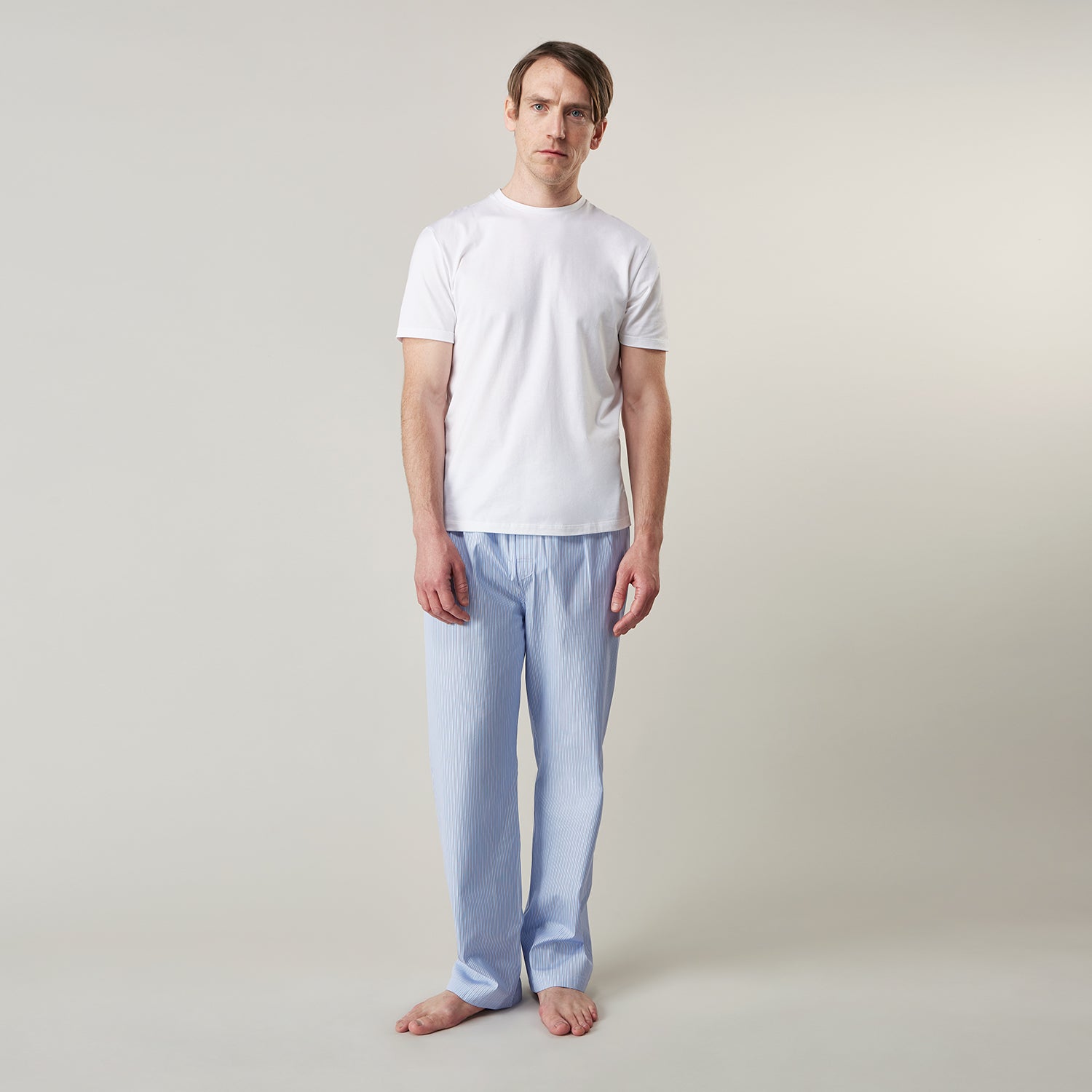 View 1 of product A man stands barefoot against a plain background, wearing a white short-sleeve T-shirt and Turnbull & Asser Blue Stripe Cotton Pyjama Trousers in a contemporary fit, looking straight ahead with a neutral expression.