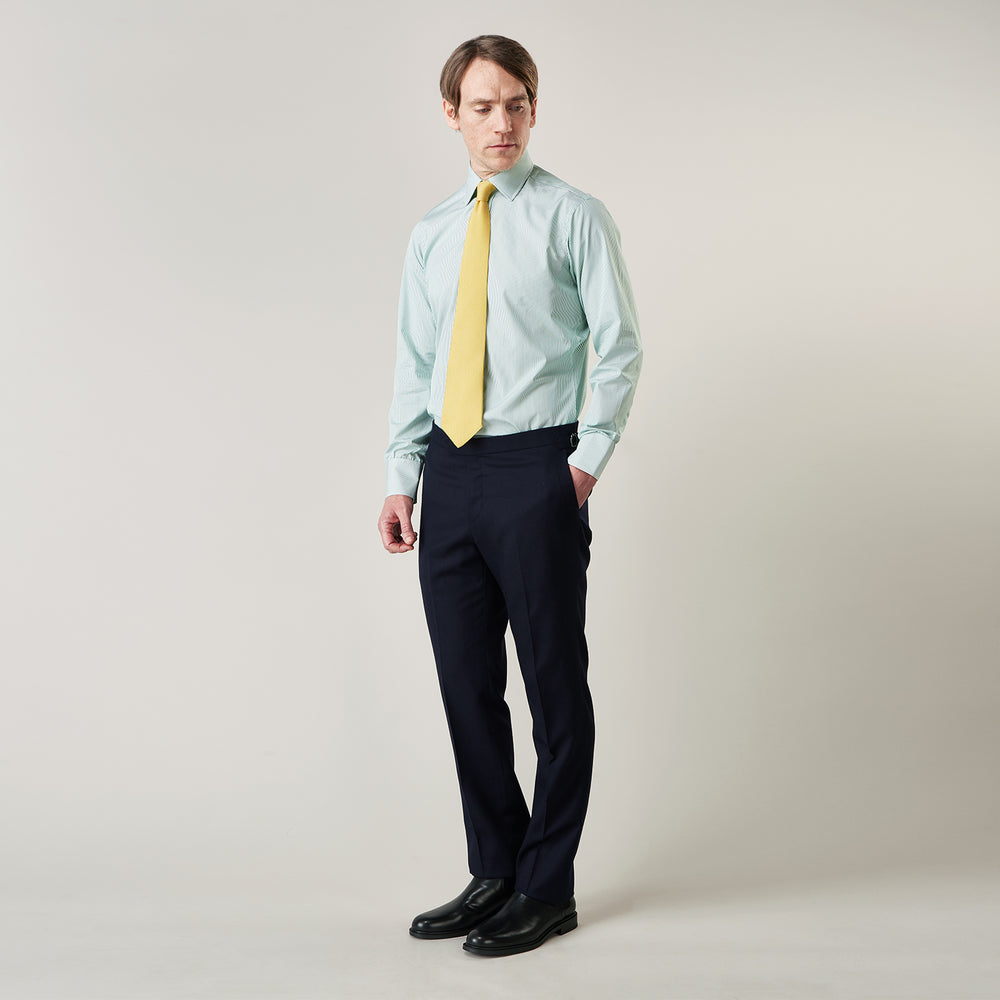 A man stands against a plain background, wearing a Turnbull & Asser Green Fine Stripe Regular Fit Shirt with a T&A collar, paired with a yellow tie, dark trousers, and black shoes. He looks slightly left with his hands at his sides.