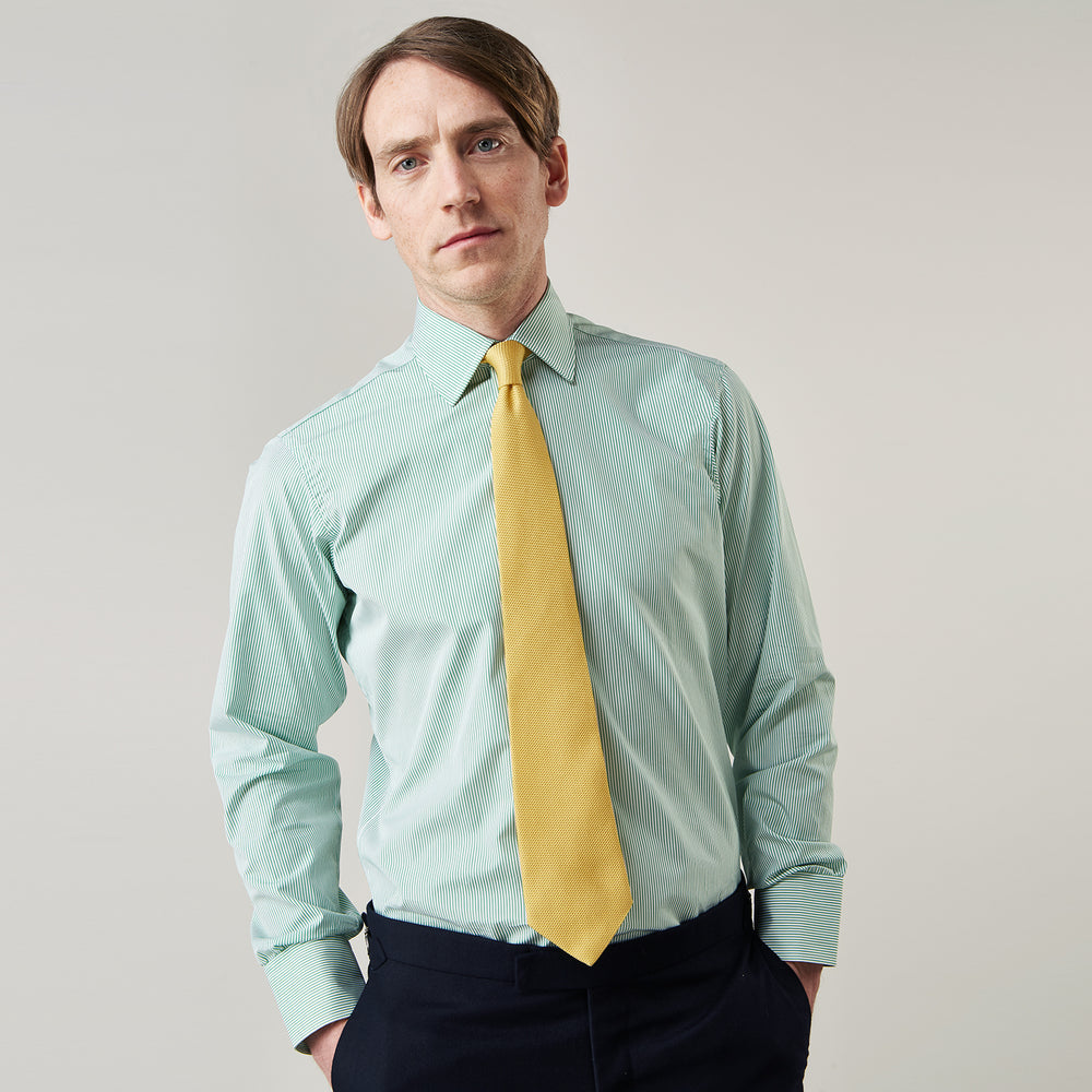 A man stands against a plain background, wearing a Turnbull & Asser Green Fine Stripe Regular Fit Shirt with a T&A collar, paired with a yellow tie, dark trousers, and black shoes. He looks slightly left with his hands at his sides.