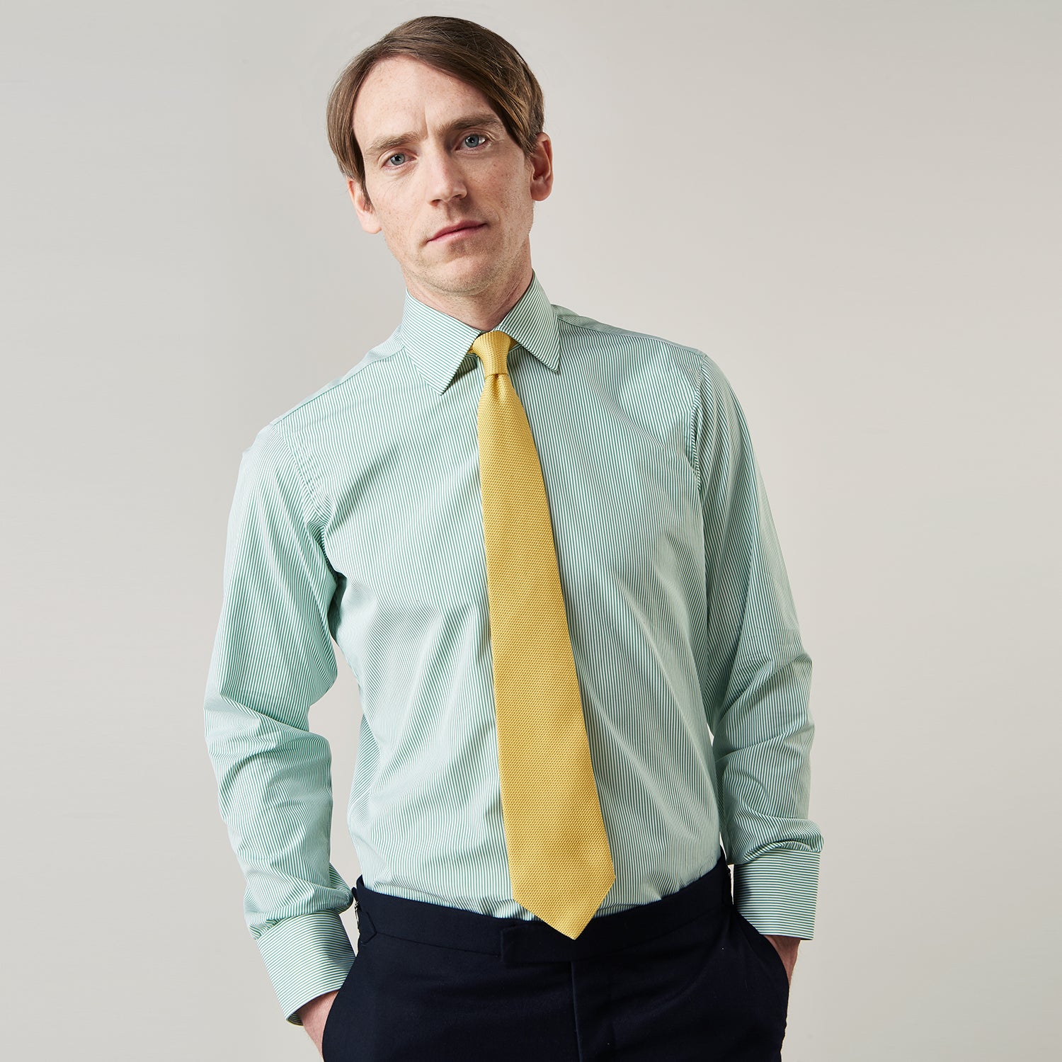 View 3 of product A man stands against a plain background, wearing a Turnbull & Asser Green Fine Stripe Regular Fit Shirt with a T&A collar, paired with a yellow tie, dark trousers, and black shoes. He looks slightly left with his hands at his sides.
