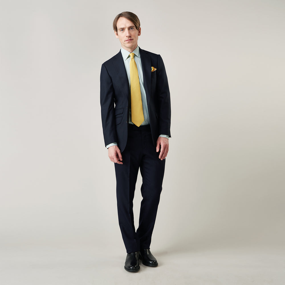 A man stands against a plain background, wearing a Turnbull & Asser Green Fine Stripe Regular Fit Shirt with a T&A collar, paired with a yellow tie, dark trousers, and black shoes. He looks slightly left with his hands at his sides.