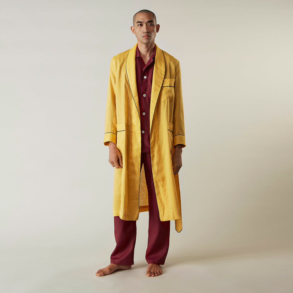 The Turnbull & Asser Ochre Linen Piped Gown features black piping on the collar, cuffs, and edges, a chest pocket, two front pockets, and a matching waist belt. Made with breathable linen for comfort, shown against a plain white background.