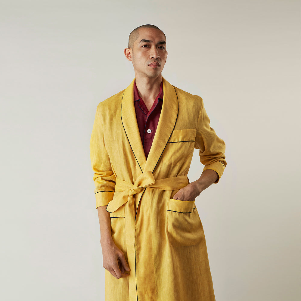 The Turnbull & Asser Ochre Linen Piped Gown features black piping on the collar, cuffs, and edges, a chest pocket, two front pockets, and a matching waist belt. Made with breathable linen for comfort, shown against a plain white background.