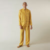 View 3 of product A barefoot person stands against a plain background, wearing the Turnbull & Asser Ochre Linen Modern Pyjama Set with black piping on the collar, cuffs, and pockets. The button-down top completes the look as they gaze to the side.