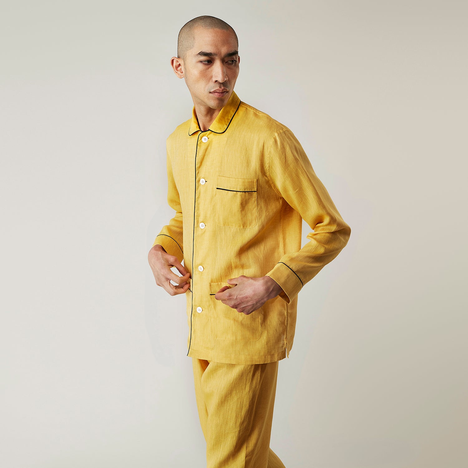View 8 of product A barefoot person stands against a plain background, wearing the Turnbull & Asser Ochre Linen Modern Pyjama Set with black piping on the collar, cuffs, and pockets. The button-down top completes the look as they gaze to the side.