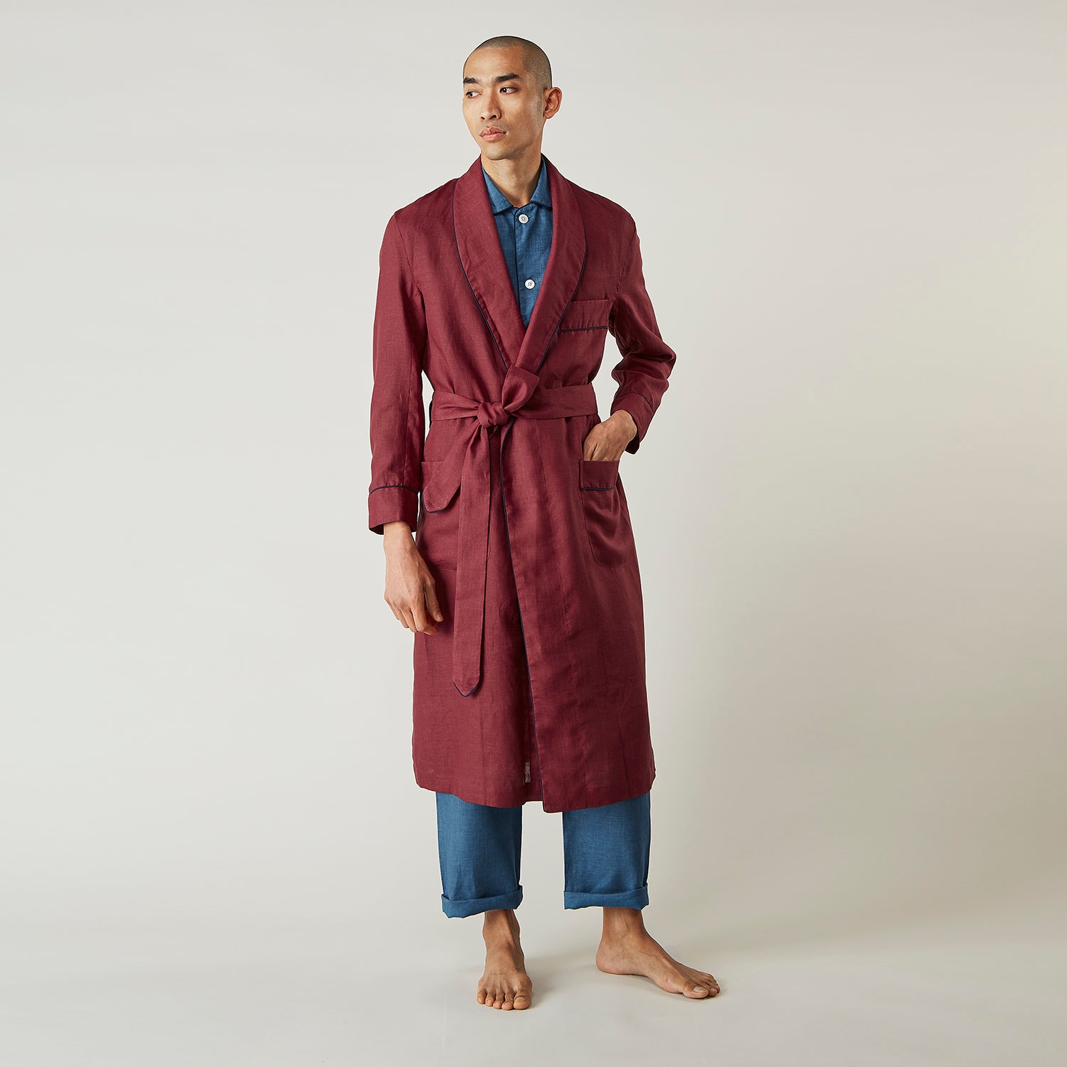 View 1 of product A person stands barefoot against a plain background, wearing the Turnbull & Asser Burgundy Linen Piped Gown belted at the waist over blue pajamas, looking to the side with one hand in the gown’s pocket.
