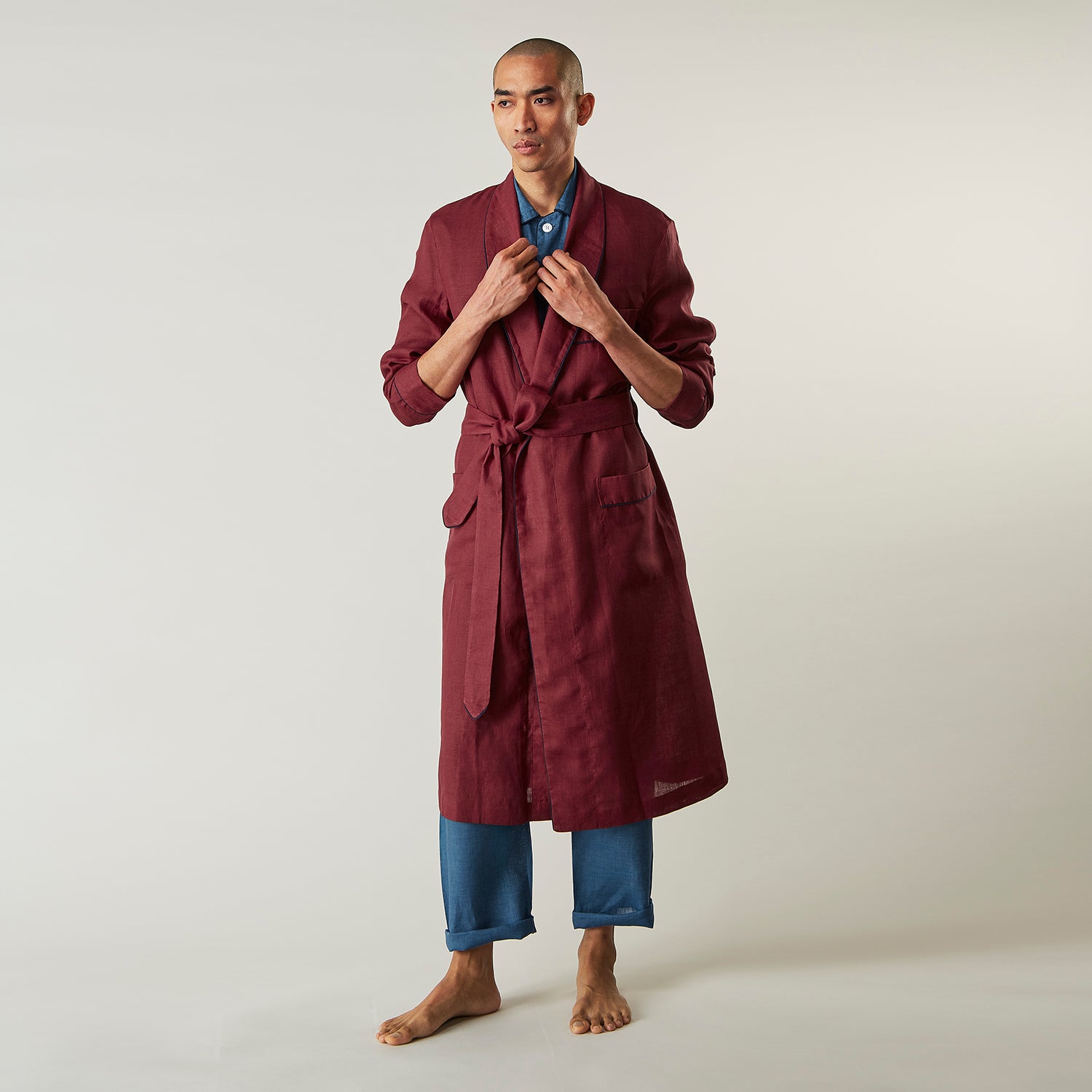 View 7 of product A person stands barefoot against a plain background, wearing the Turnbull & Asser Burgundy Linen Piped Gown belted at the waist over blue pajamas, looking to the side with one hand in the gown’s pocket.