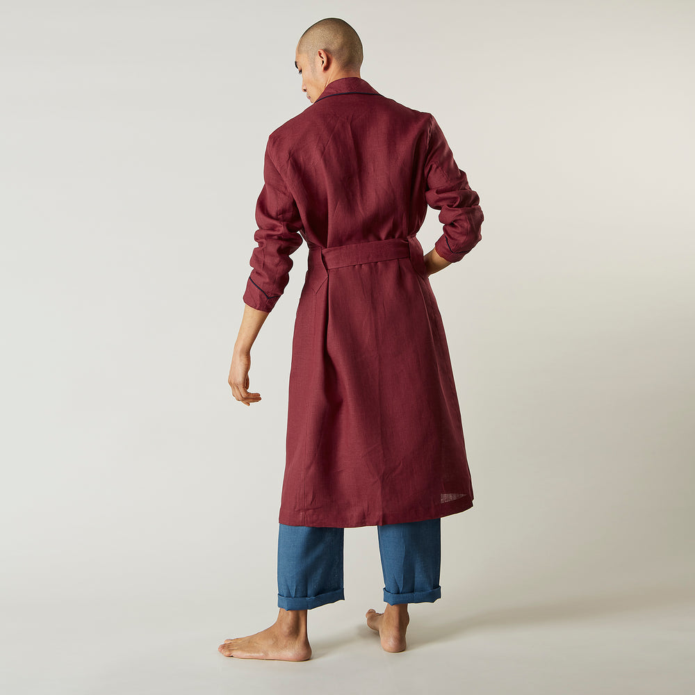 A person stands barefoot against a plain background, wearing the Turnbull & Asser Burgundy Linen Piped Gown belted at the waist over blue pajamas, looking to the side with one hand in the gown’s pocket.