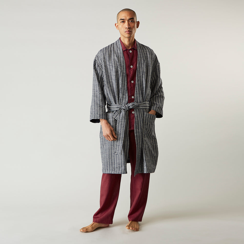 A barefoot person wears Turnbull & Asser's Navy Silk Chequered Kimono Gown with a belt over maroon pyjama sets, standing against a plain light background.