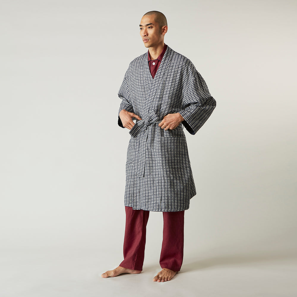 A barefoot person wears Turnbull & Asser's Navy Silk Chequered Kimono Gown with a belt over maroon pyjama sets, standing against a plain light background.