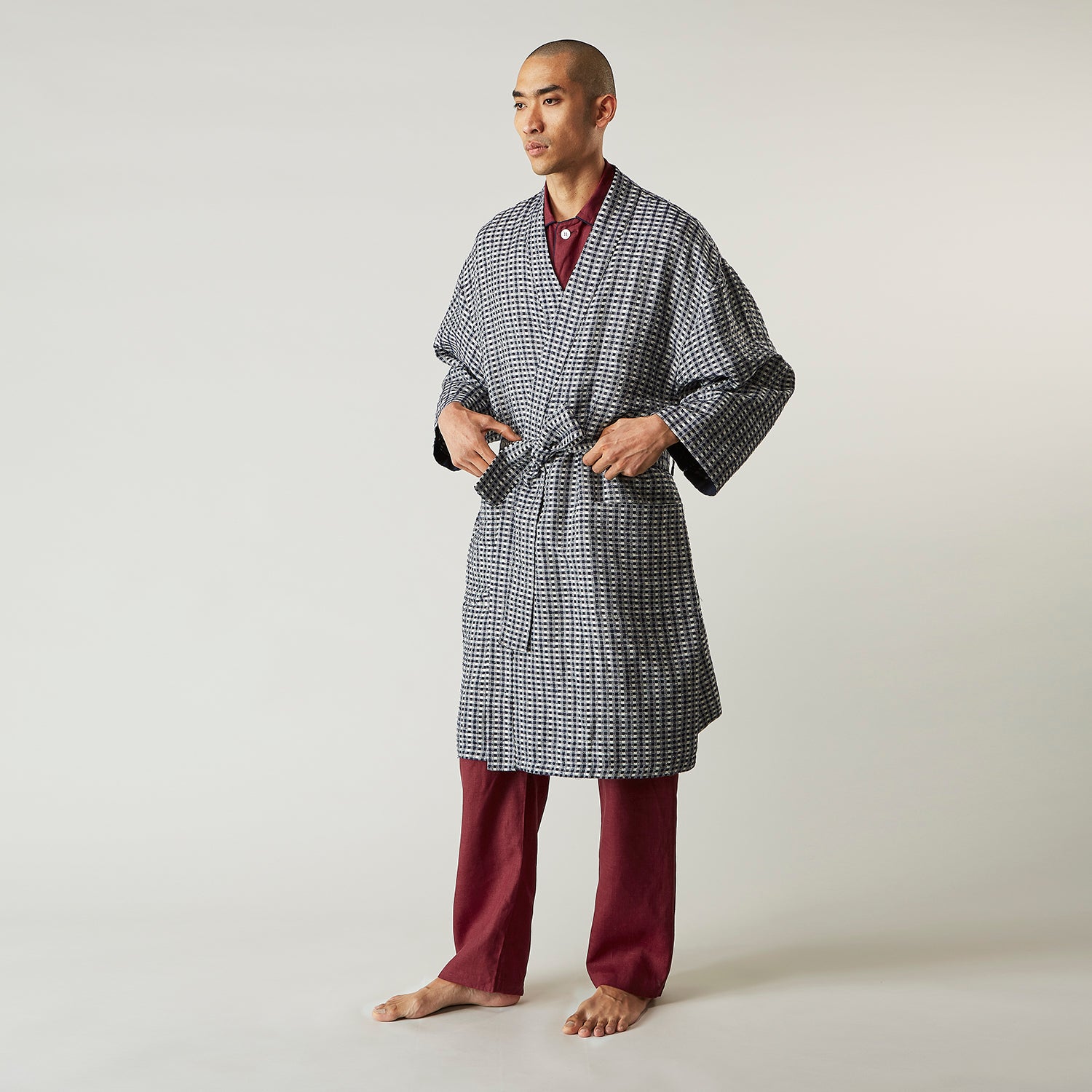 View 3 of product A barefoot person wears Turnbull & Asser's Navy Silk Chequered Kimono Gown with a belt over maroon pyjama sets, standing against a plain light background.