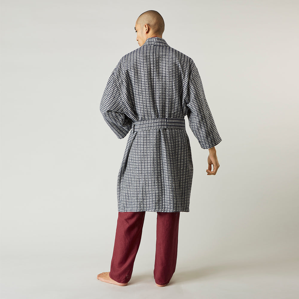 A barefoot person wears Turnbull & Asser's Navy Silk Chequered Kimono Gown with a belt over maroon pyjama sets, standing against a plain light background.