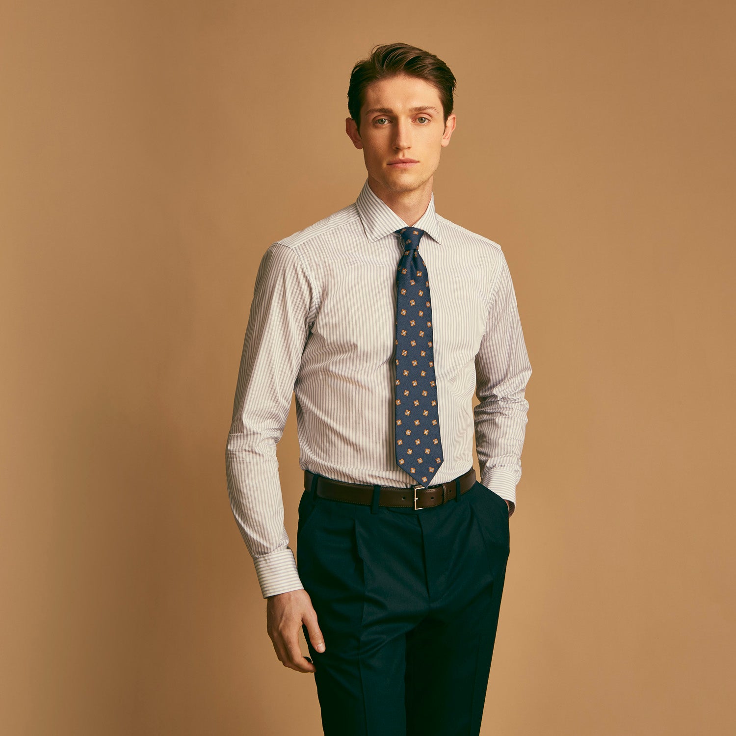 View 1 of product A man wearing the Turnbull & Asser Grey Multi Stripe Regular Fit Twill Shirt with T&A Collar and Double Cuffs stands against a beige background, one hand in his pocket, looking at the camera.