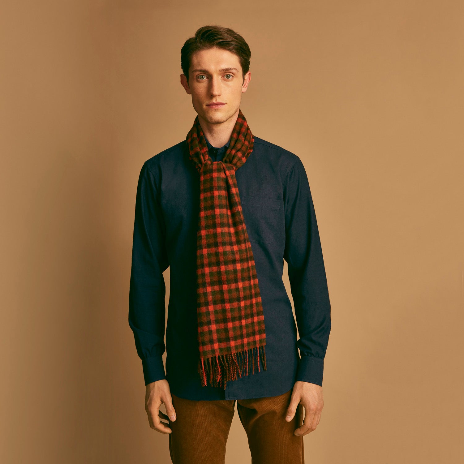View 1 of product A man stands against a beige background wearing a navy long-sleeve shirt, brown pants, and a Turnbull & Asser Orange Multi Shepherd Check Cashmere Scarf draped around his neck. He faces forward with a neutral expression.
