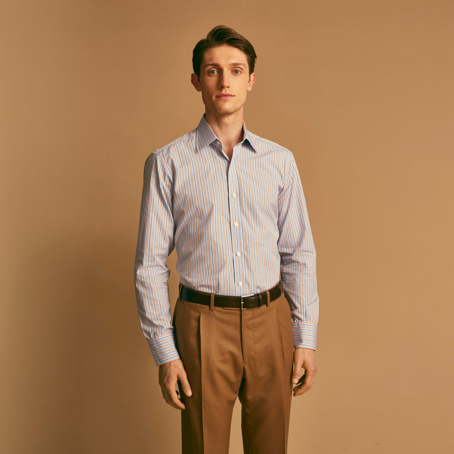 View 1 of product A man stands against a beige background, wearing a Turnbull & Asser Blue, Orange & Navy Multi Stripe Regular Fit Shirt with T&A collar and 3-button cuffs, tucked into tan dress pants with a black belt. He faces forward with a neutral expression.