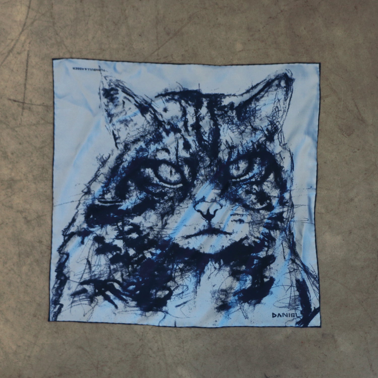 View 2 of product The Turnbull & Asser DANIEL PERKIN BLUE WILDCAT POCKET SQUARE features a blue-toned sketch of a tabby cat’s face with vivid eyes and fine fur detail on a pale blue background, finished with “DANISL” in the lower right corner.