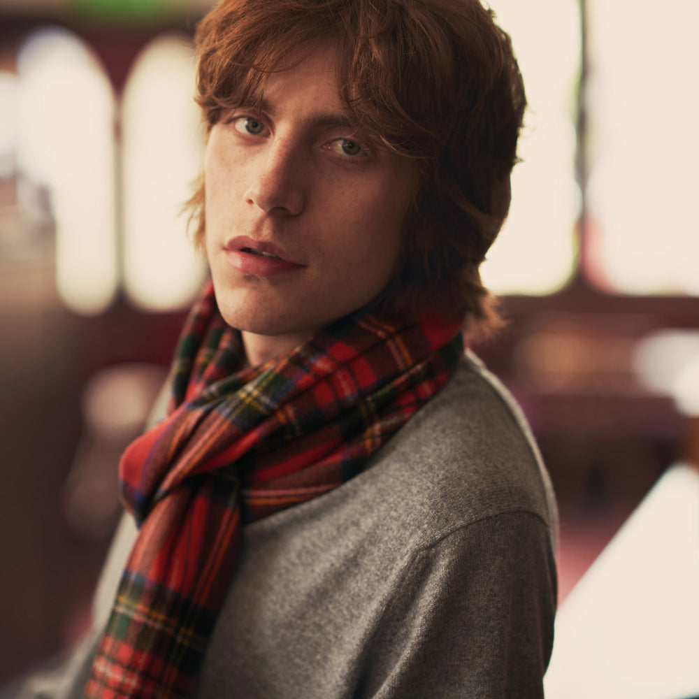 The Turnbull & Asser Royal Stewart Tartan Cashmere Scarf, showcasing a classic red plaid with black, yellow, and green accents and fringed ends, is displayed against a white background.