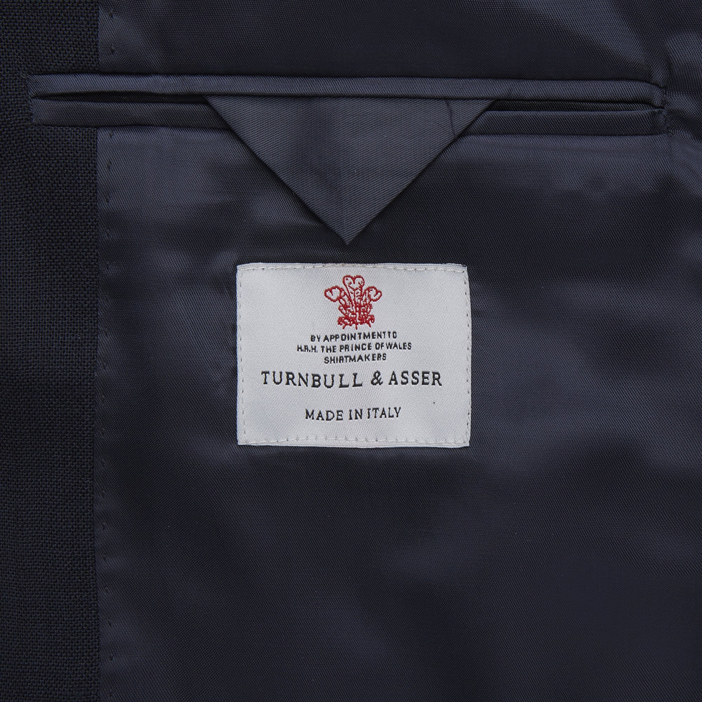 The Turnbull & Asser Navy Lightweight Wool Hopsack Traveller Jacket features a notched lapel, three front buttons, and four flap pockets—two on the chest and two at the waist—all displayed on a plain white background.