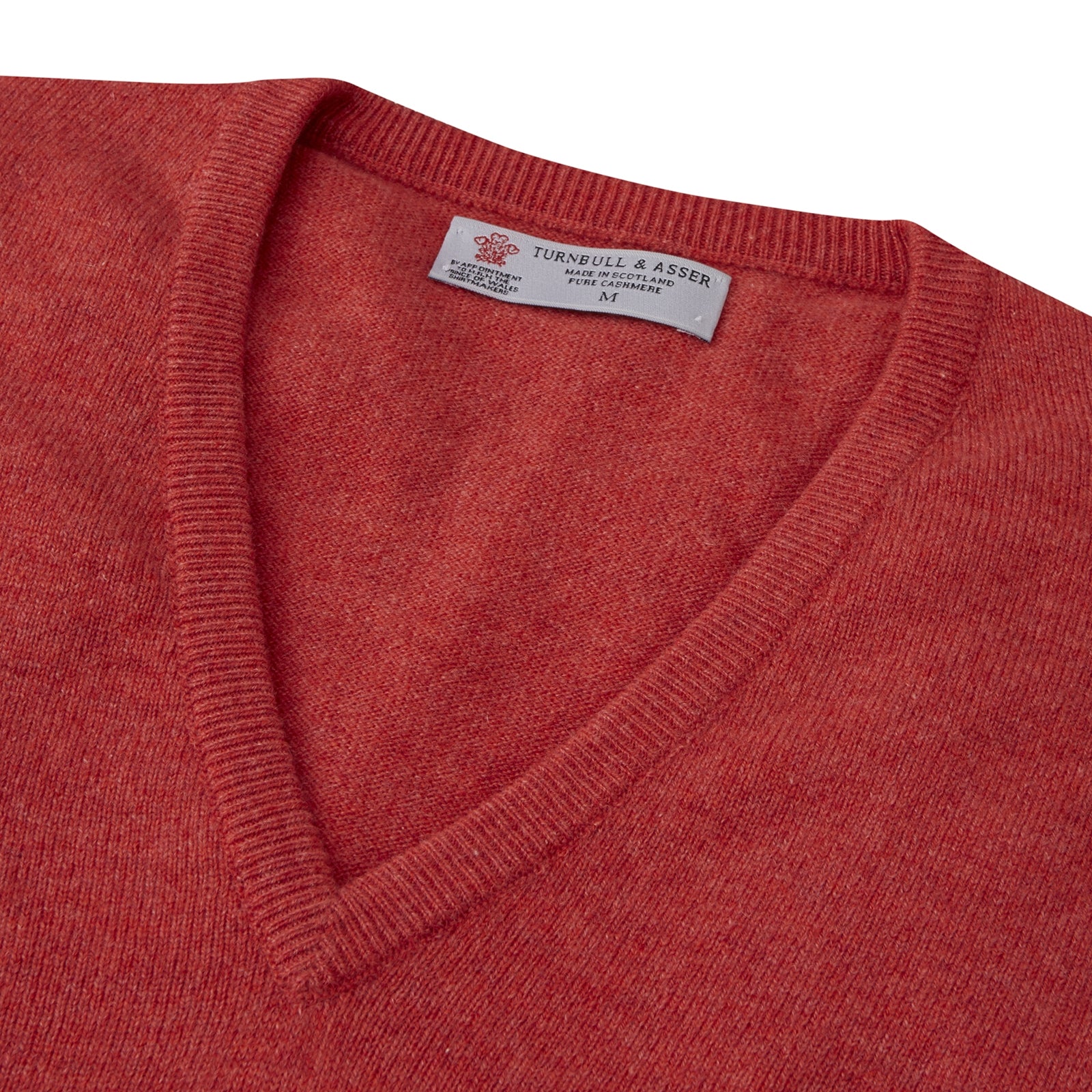 View 2 of product A folded Crimson Fine Cashmere Slipover by Turnbull & Asser in coral-red is shown against a white background, with the visible neckline label highlighting its premium Scottish cashmere quality.