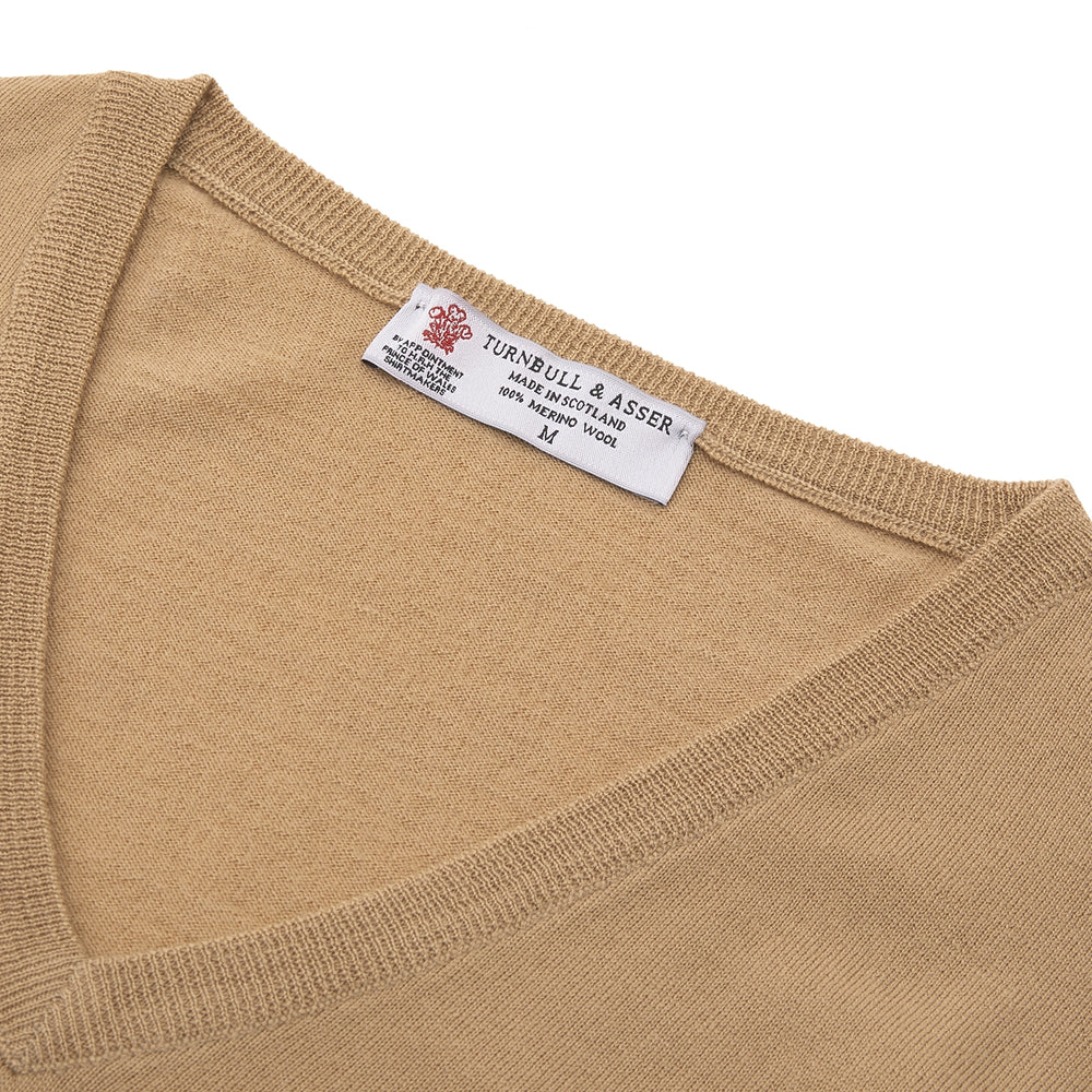 The Turnbull & Asser Camel V-Neck Merino Wool Jumper, with ribbed cuffs and a visible white neckline label, is neatly folded with one sleeve showing—an essential, versatile staple for any gentleman's wardrobe.