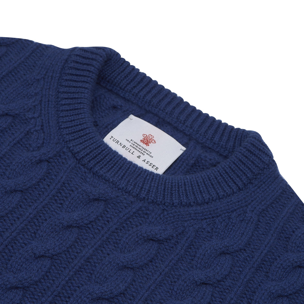 The Blue Crew Neck Cable Knit Cashmere Jumper by Turnbull & Asser features a chunky cable knit, ribbed crew neck and cuffs, and a visible label inside the collar.