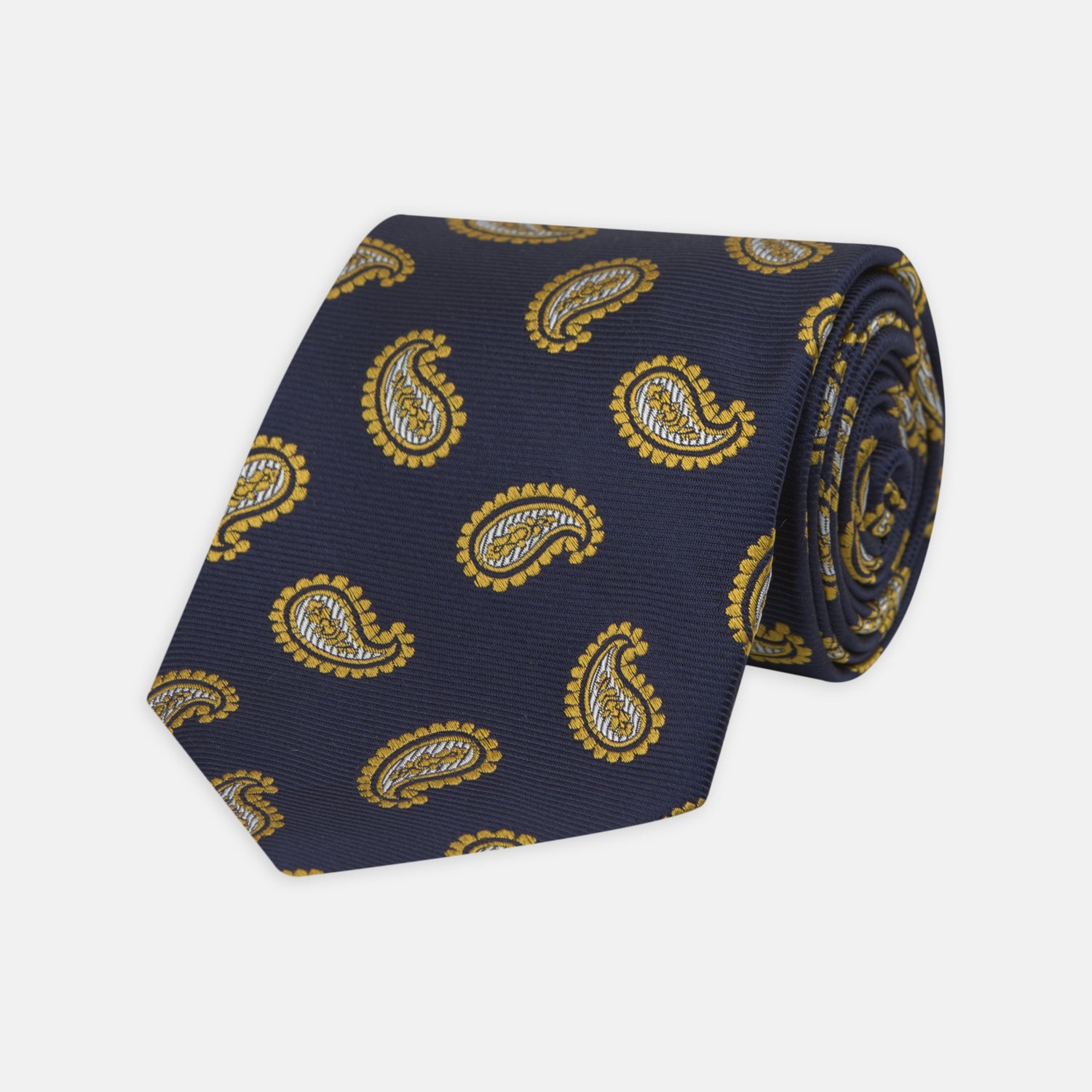 View 1 of product The Turnbull & Asser Navy and Gold Floating Paisley Silk Tie features a repeating yellow and white paisley pattern, shown rolled with the pointed end facing forward against a white background.