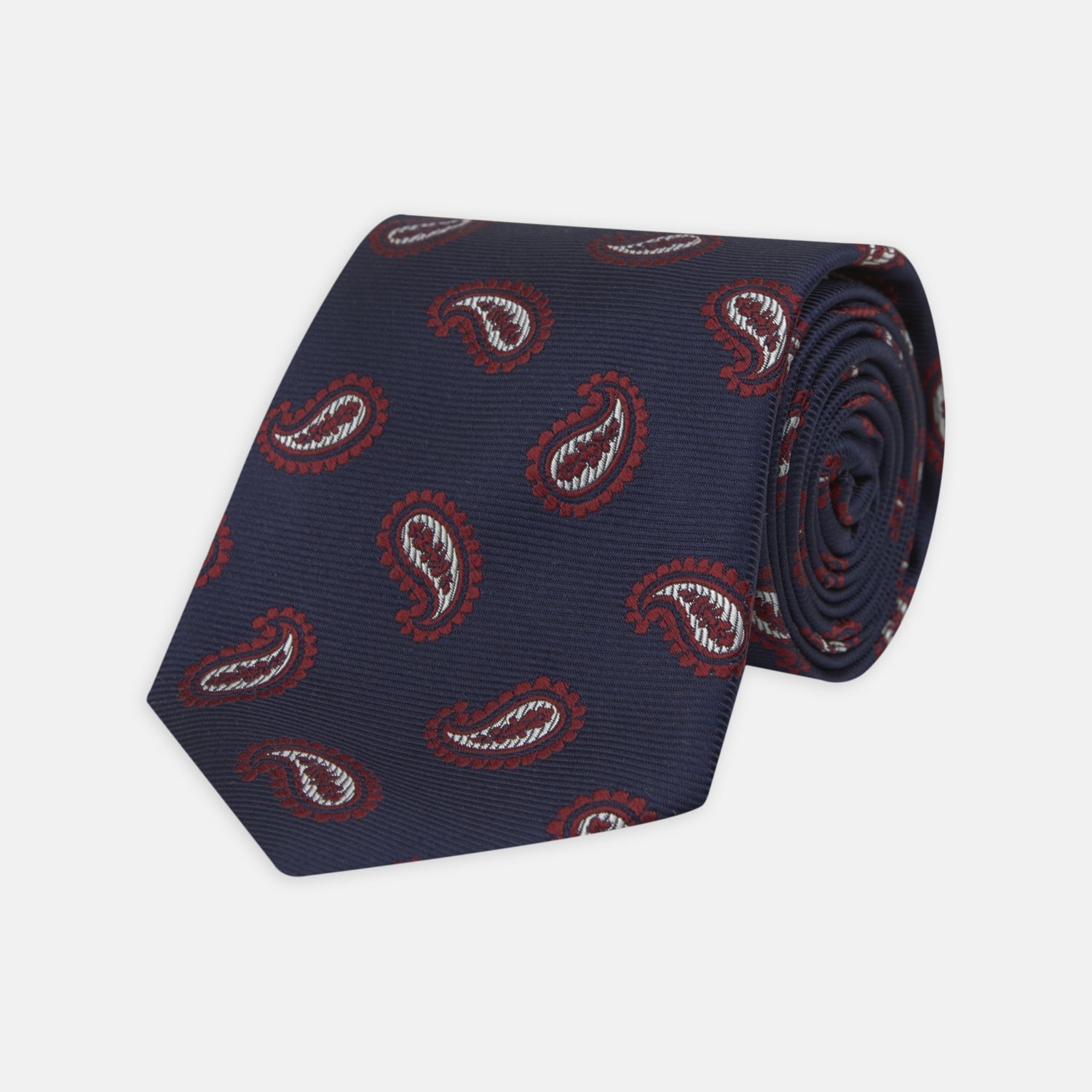 View 1 of product The Turnbull & Asser Navy and Burgundy Floating Paisley Silk Tie is shown rolled up, displaying its repeating paisley design on a plain white background.