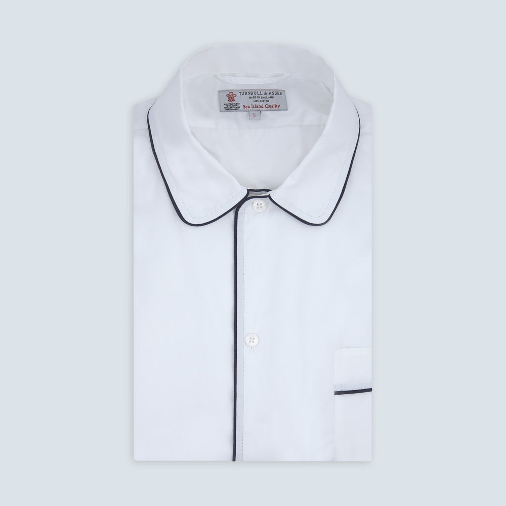 The Turnbull & Asser White Sea Island Quality Cotton Nightshirt, featuring black piping on the collar and pocket, is elegantly displayed folded against a light grey background.