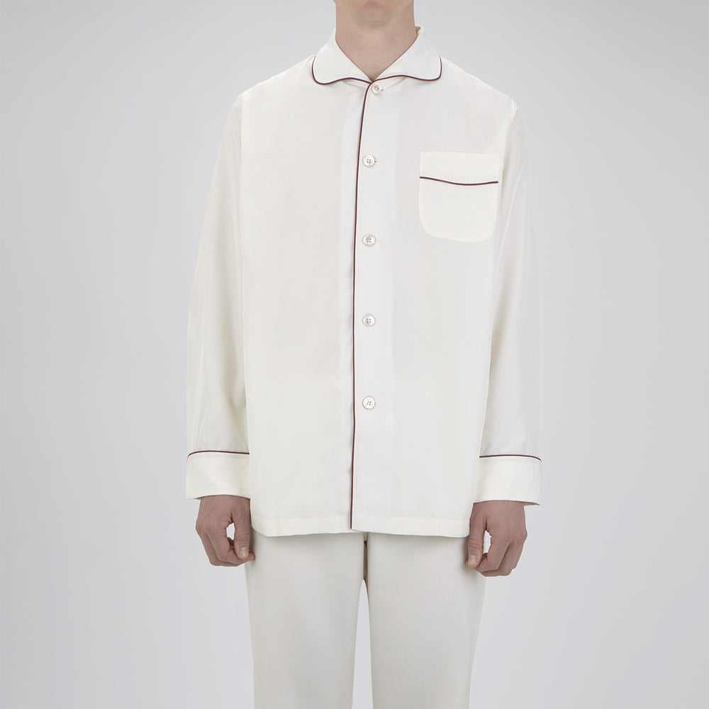 The Turnbull & Asser Cream Piped Silk Pyjama Set is neatly folded with dark piping and buttons, placed next to a matching white sleep mask with tie straps, all set against a light neutral background.