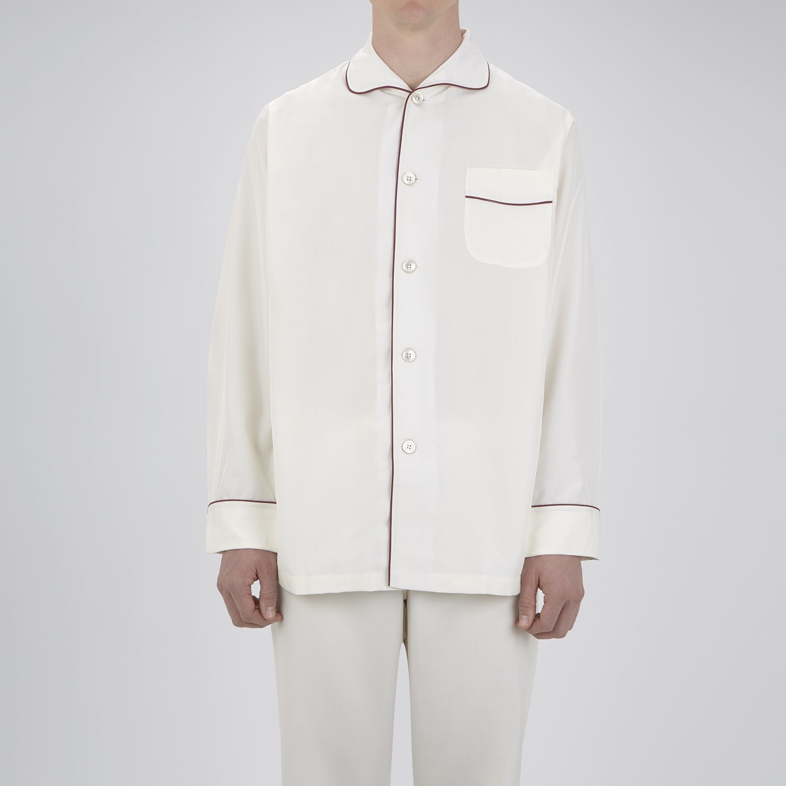 View 3 of product The Turnbull & Asser Cream Piped Silk Pyjama Set is neatly folded with dark piping and buttons, placed next to a matching white sleep mask with tie straps, all set against a light neutral background.